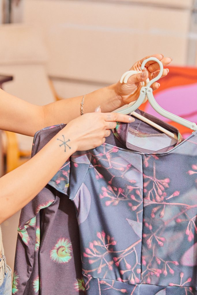 Image: Marissa is hanging a top on a rack where several other tops hang. The tops are mostly pale purple. Photo by Sarah Joyce.