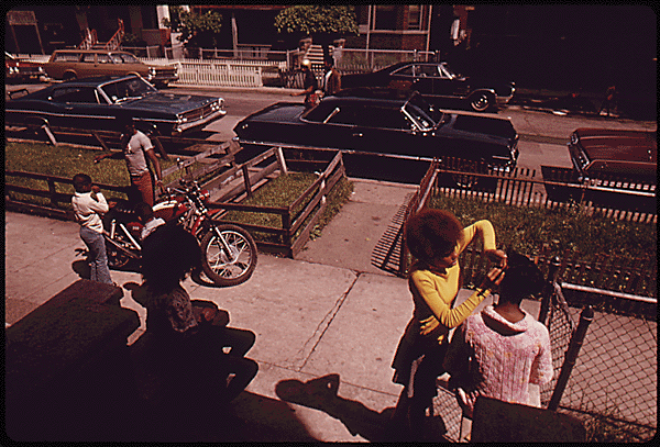 Black neighbors spending time outside on a sunny day on Chicago's West Side in 1974. On the left, two children stand together, one holding a bike. In the shadow of the home that falls outside of the frame, another child sits on the porch. To the right, two young people stand, one with their hands in the hair of the other, braiding. Cars line the street in front of them. Photo from John H. White's series DOCUMERICA: The Environmental Protection Agency's Program to Photographically Document Subjects of Environmental Concern, 1972 - 1977. Source: The National Archives and Records Administration.