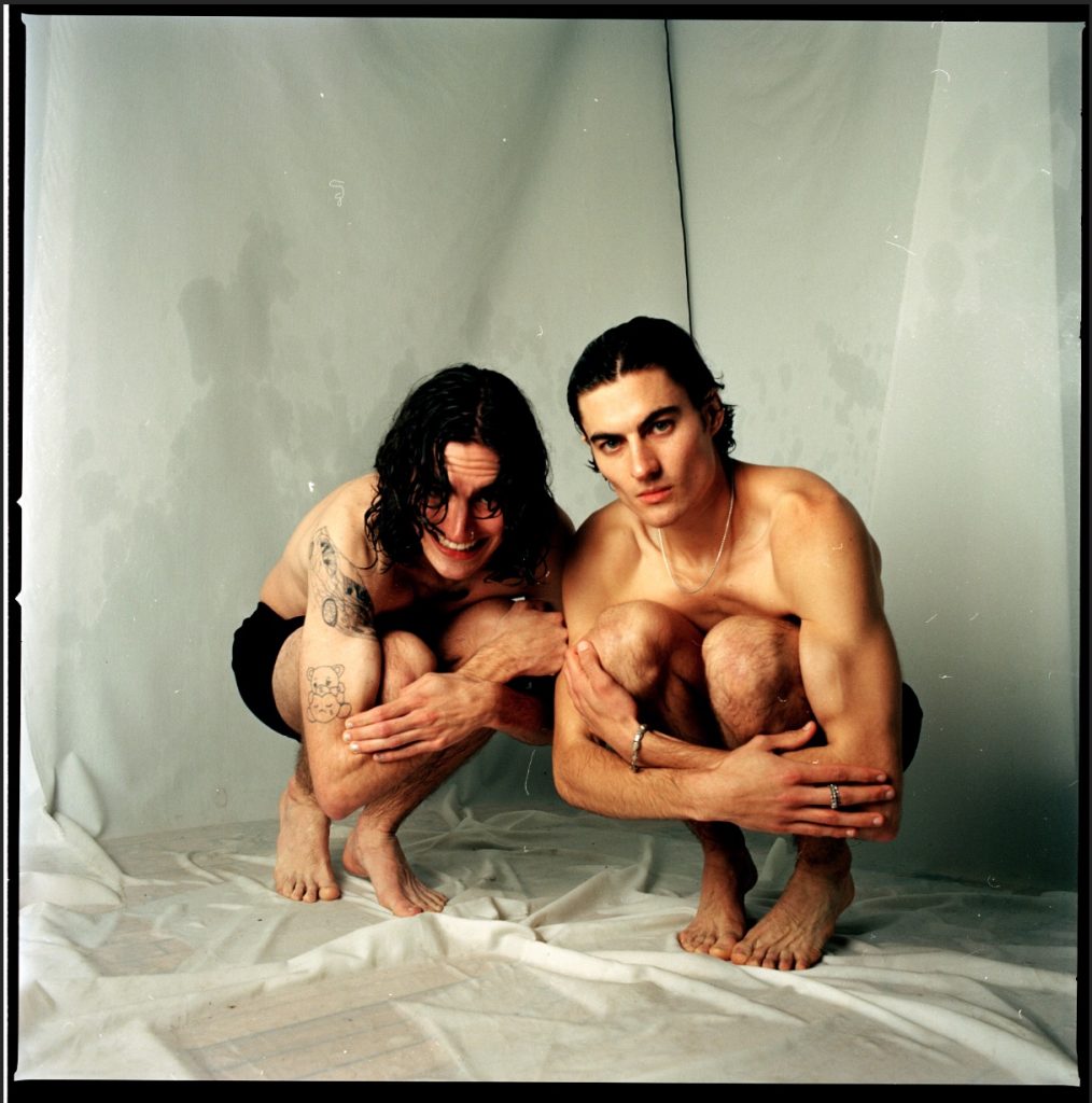Image:  A photo of boys crouching down in the corner of a wet studio, holding themselves as if they just got out of a lake and they’re warming themselves up. The boys both look directly into the camera, one serious and one smiling with hair in front of their eyes. Title: Will and John. Year: 2021. Image by Madeline Hampton