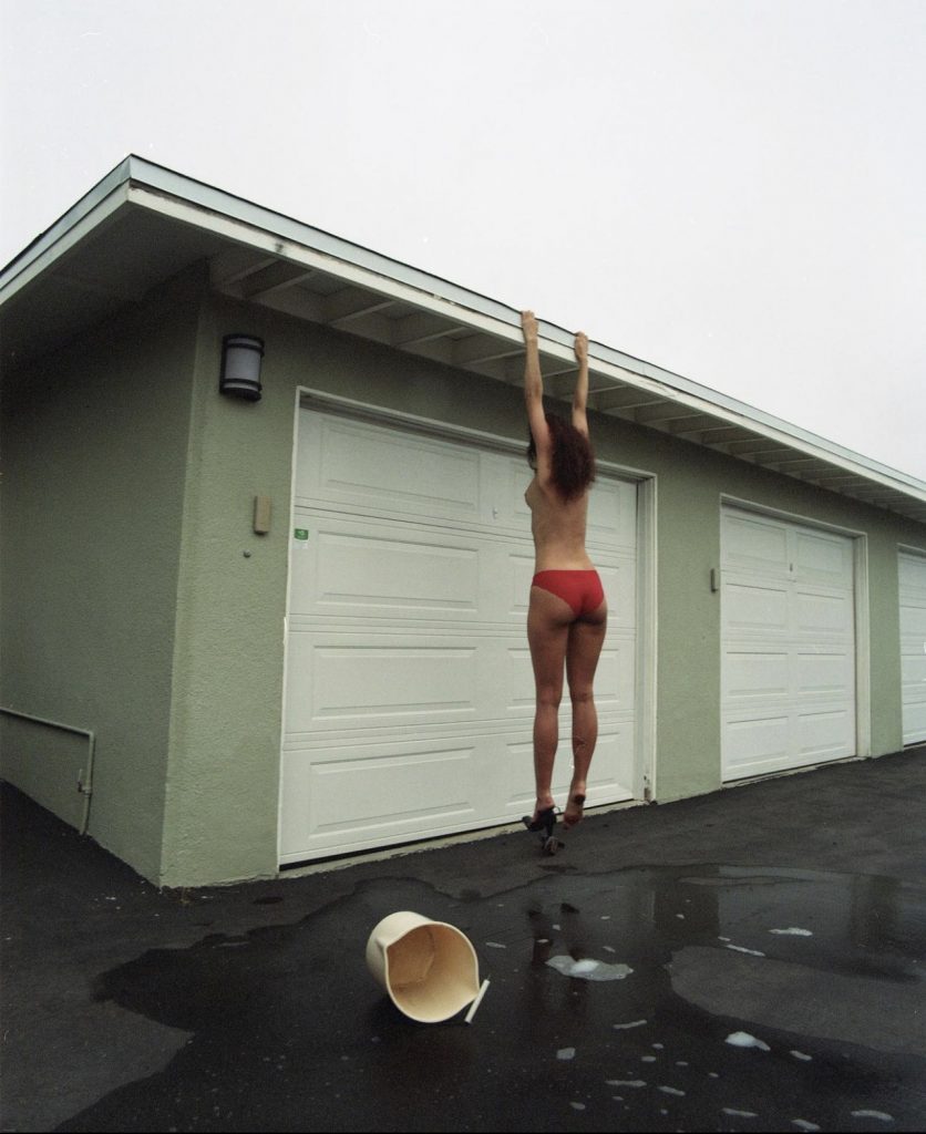 Image: A white woman with brown hair wearing red underwear and hanging off the side of a green garage (as if about to do a pull-up). A bucket has fallen down and there is soapy water streaming over the black tarmac. Title:  Likeapendulum. Year: 2020. Image: Madeline Hampton. 