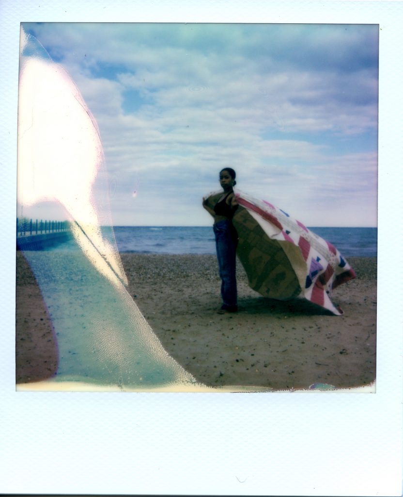 Image: A Polaroid photograph of Rikki Byrd standing on a beach in front of Lake Michigan with her body facing the left side of the photograph as she looks forward at the viewer. She holds a quilt made by her great grandmother around her shoulders like a cape while it blows in the wind. There is slight bluish coloring on the left side of the Polaroid. Jeans are vintage Levi’s. Styling by row särkelä and Madeleine Le Cesne. Photo by Jared Brown. 