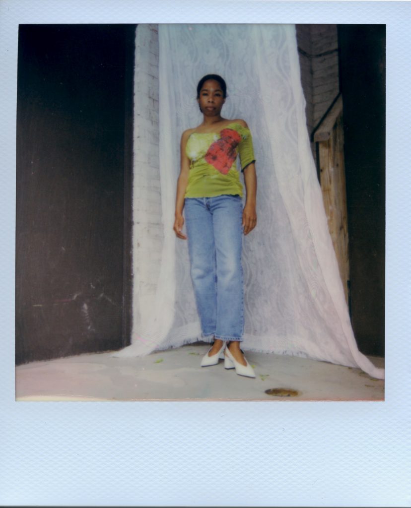 Image: A Polaroid photograph of Rikki Byrd wearing vintage blue Levi’s jeans, white heels from Zara, and a greenish-yellow handmade corset top from @earthangelbaby on Depop with one sleeve and a red patch. Draped behind her is white lace. She stands while looking forward at the viewer. Styling by row särkelä and Madeleine Le Cesne. Photo by Jared Brown.