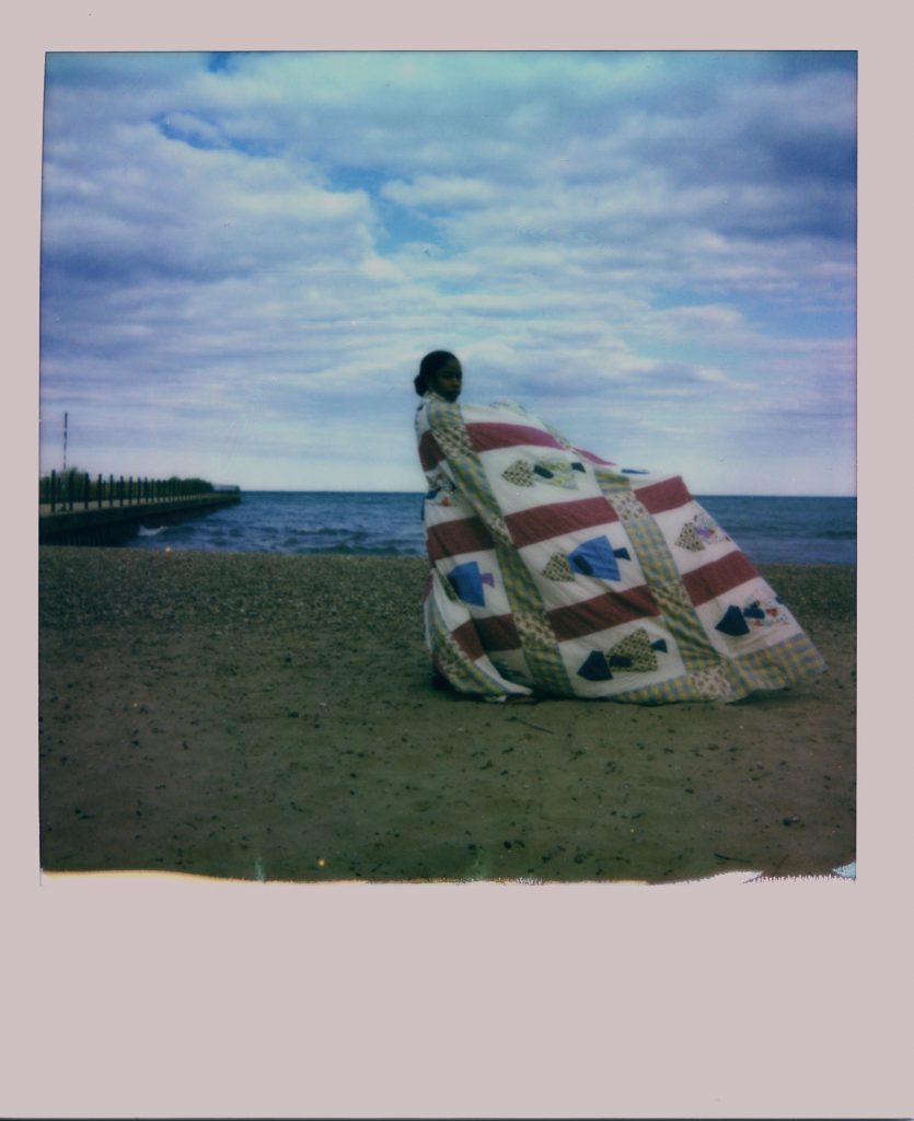 Image: A Polaroid photograph of Rikki Byrd standing on a beach in front of Lake Michigan and is wrapped in a quilt made from women in her family. She look directly at the viewer while the quilt blows in the wind. Photo by Jared Brown.