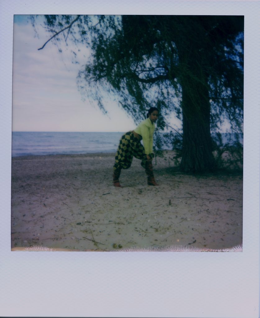 Image: A Polaroid photograph of Rikki Byrd standing on a beach while bending to the right with her hands on one knee. She wears yellow and black checkered pants, boots, and a yellow top while looking at the viewer. Photo by Jared Brown.