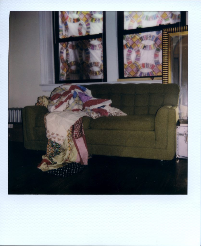 Image: A Polaroid photograph of a green couch with a quilt laying on it in Rikki Byrd’s apartment. There are patches of patterned quilt pieces in the background covering the windows like a kaleidoscope. Photo by Jared Brown. 