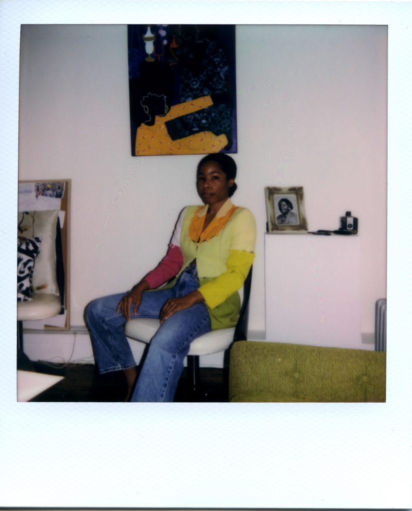 Image: A Polaroid photograph of Rikki Byrd sitting on a white chair in her apartment. She wears a blue jeans and a shirt that is magenta, yellow, green, white, and orange. Next to her sits a photo of her great-grandmother and a camera. She has her hands in her lap and looks directly at the viewer. Photo by Jared Brown.