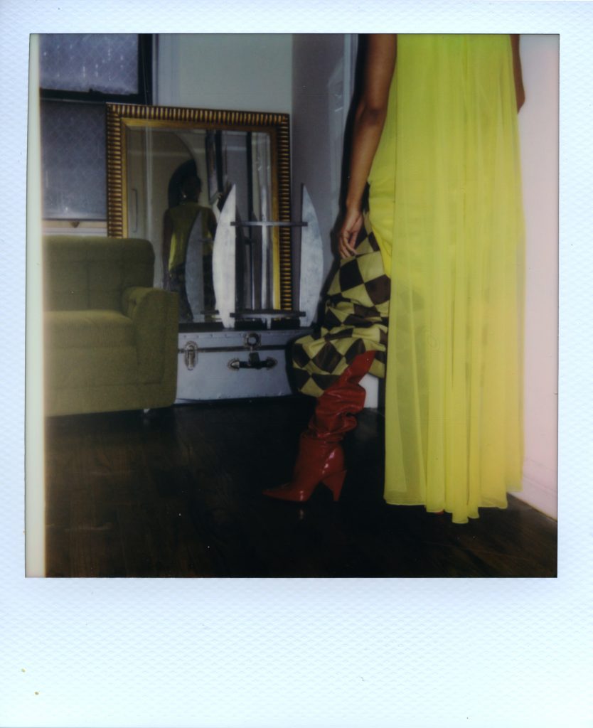 Image: A Polaroid photograph of Rikki Byrd standing in front of a mirror that is next to her green couch. The foreground shows Rikki from the back wearing red boots from Zara, green and black checkered pants from Vaquera, and a long, green dress from May’s place, a vintage shop in St. Louis. The mirror shows Rikki’s front. Styling by row särkelä and Madeleine Le Cesne. Photo by Jared Brown.