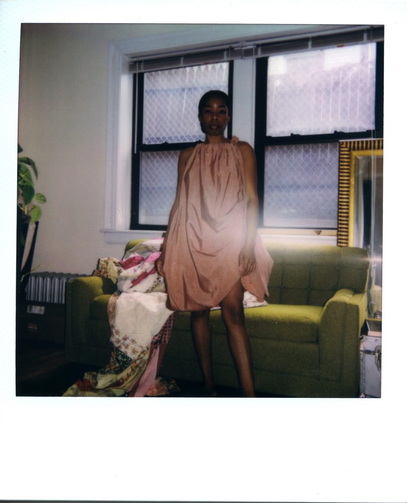 Image: A Polaroid photograph of Rikki Byrd standing in front of a window and a green couch in her apartment. A quilt lays on the couch and a mirror sits right behind the couch to the right. Rikki stands and looks straight out at the views while wearing a pale pinkish dress by Chicago-based designer Adreain Guillory. Styling by row särkelä and Madeleine Le Cesne. Photo by Jared Brown.