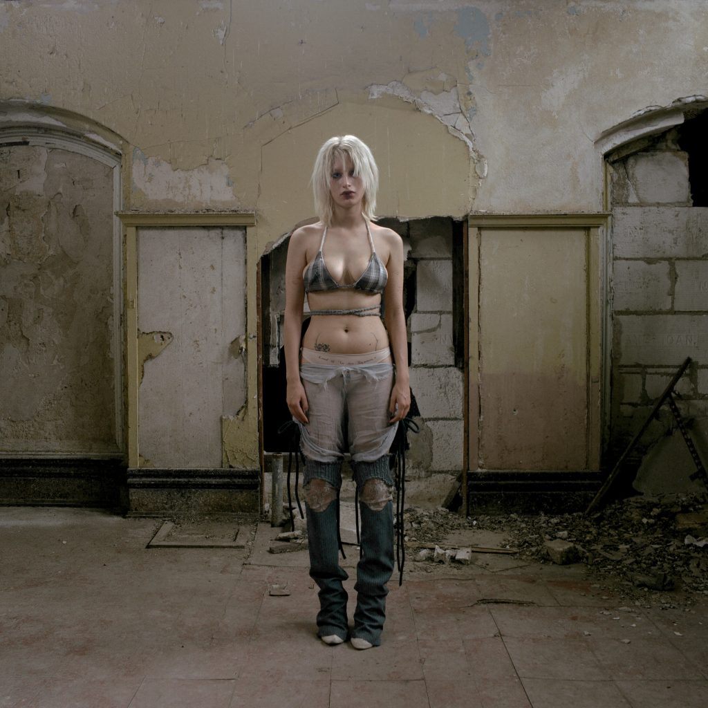 Image: A white person with white blond hair wearing deconstructed skirt, legwarmers, and bra in abandoned building. The light is yellowish and a bit eerie. Title: Genesis Lookbook for Exaltus. Year: 2021. Designer: @3xalted1. Image by Madeline Hampton. 