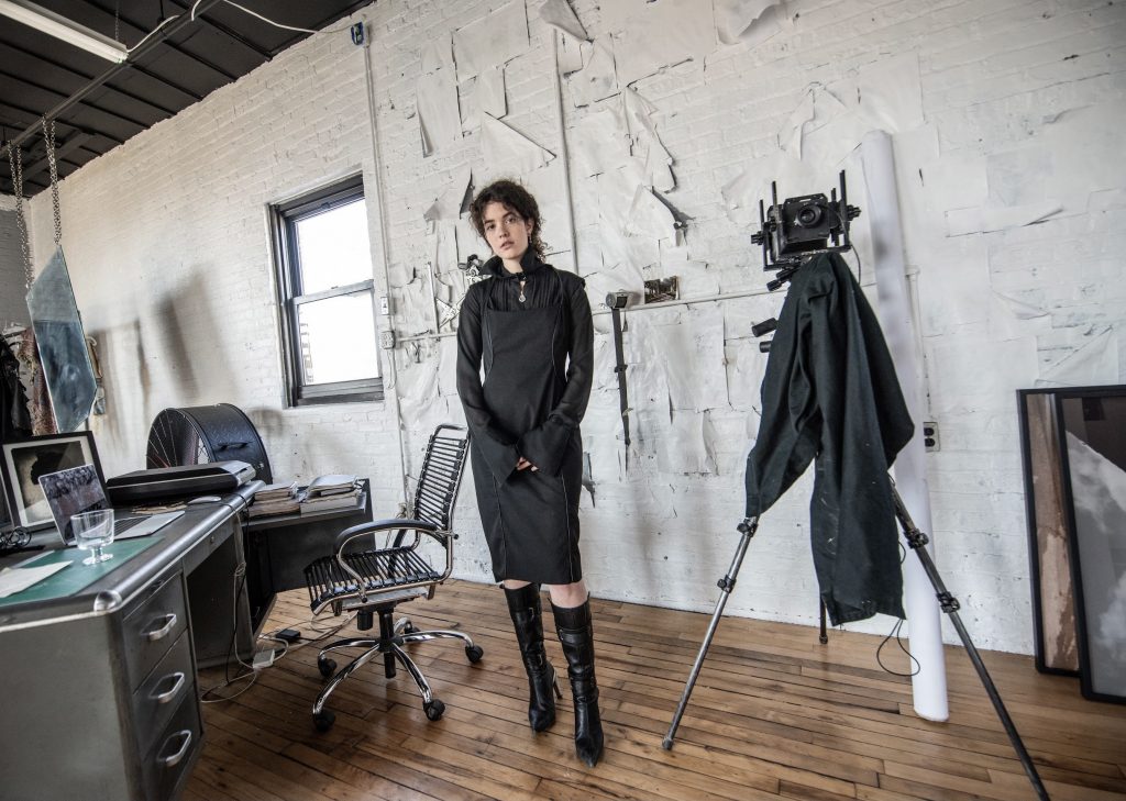 Image: A photo of Madeline Hampton in her apartment/studio. She wears a black dress and black boots and looks forward at the viewer. On the lefthand side of the photo is a window and a desk with various items sitting on top. On the righthand side is a camera on a tripod. Photo by Edvette Wilson-Jones.