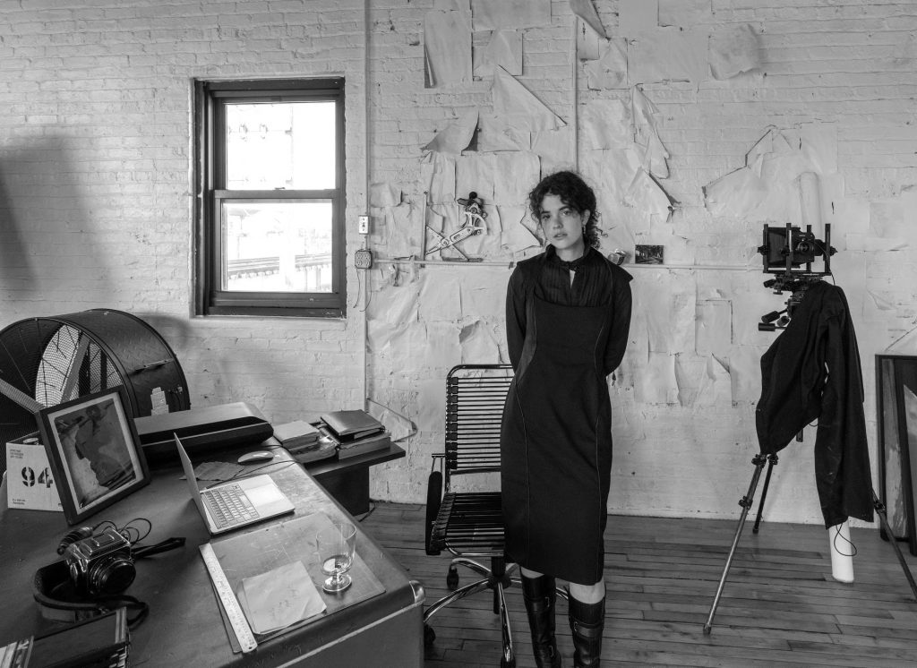 Image: A black and white photo of Madeline Hampton in her apartment/studio. She wears a black dress and black boots and looks forward at the viewer. On the lefthand side of the photo is a window and a desk with various items sitting on top. On the righthand side is a camera on a tripod. Photo by Edvette Wilson-Jones.