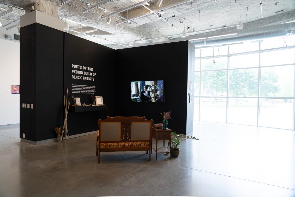 Image: An installation view of bright room with a free-standing wall at the center. The wall has a shelf holding an altar below white lettering on a black wall, and a large monitor on the adjacent wall. Just in front of the wall is a wooden upholstered seat, end table, and plants. Photo by Jessica Bingham. Courtesy of University Galleries of Illinois State University.