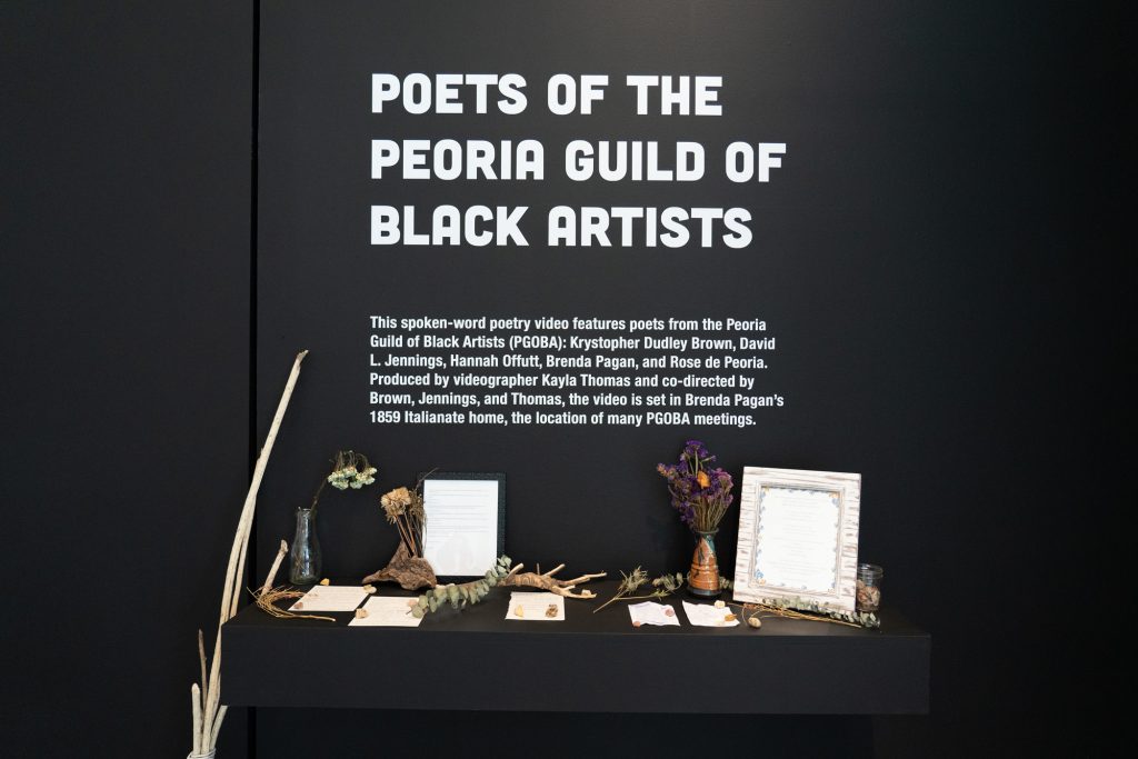 Image: An installation view of an altar of framed and loose paper resting on a shelf with dried flowers and vessels placed throughout. Above the shelf is white vinyl lettering on a black wall that reads Poets of the Peoria Guild of Black Artists with the names Krystopher Dudley Brown, David L. Jennings, Hannah Offutt, Brenda Pagan, Rose de Peoria, and Kayla Thomas underneath. Photo by Jessica Bingham. Courtesy of University Galleries of Illinois State University.