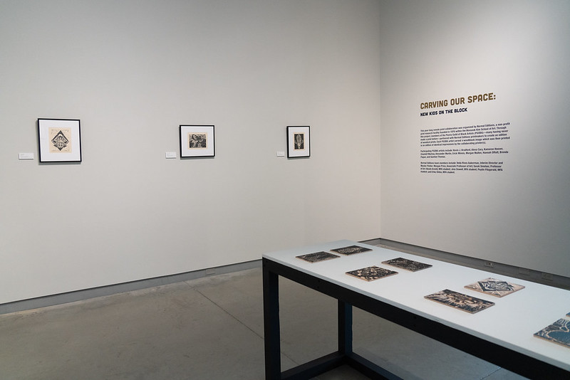 Image: Installation view of Carving Our Space: New Kids on the Block, a portfolio project organized in collaboration with Normal Editions. The image shows three framed one-color woodcut prints on Fuji Okawara paper with a table displaying the woodblocks that these pieces and others were printed on. The adjacent wall has vinyl lettering, the largest reading "Carving Our Space." Photo by Jessica Bingham. Courtesy of University Galleries of Illinois State University.