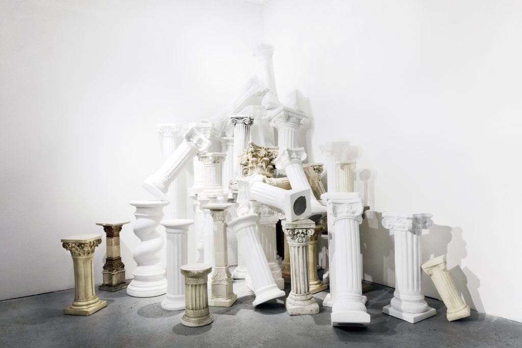 Image: Kelly Kristin Jones, "Orders of Empire," 2020 - ongoing. Plaster, wood, concrete, fiberglass composite, polyethylene, resin. The installation is a pile of plastic, white, faux columns in the corner of 062 Gallery. Image courtesy of the artist.