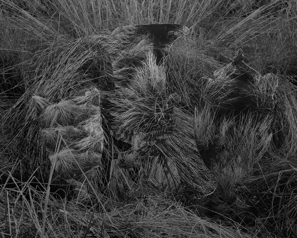 Image: Kelly Kristin Jones, "Untitled (Grave goods)," 2021. Archival pigment print, 16in x 20in. Edition of 3. A black and white photograph of long, unkempt grass. Three ghostly shapes of vases are sitting on the ground. Image courtesy of the artist.