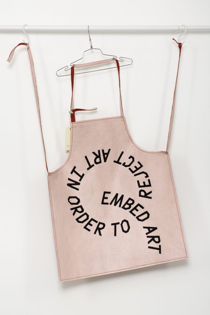 Image: An apron in pink felt by Nat Pyper gives protest-banner-energy and spells out REJECT ART IN ORDER TO EMBED ART in a big spiral of black block letters across the whole front. Photo courtesy of LVL3.