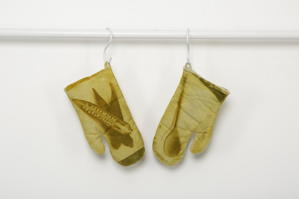 Image: Two oven mitts in a hazy yellow by Chicago-based artist Janelle Ayana Miller, titled “Bless the Hands that Prepare.” The right glove features a printed photo transfer of a wooden spoon, and the left glove shows a printed photo transfer of the open side of an okra cut longwise. Both transfers are in darker yellow, mixed with an olive green, against the lighter yellow of the mitt. The photos honor Miller’s family's culinary practices and maternal figures. Photo courtesy of LVL3.