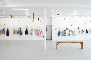 Featured image: Overview of LtdWear5 installed at the bright LVL3 gallery. The image is a landscape shot of about thirty colorful aprons and oven mitts displayed on racks on the far white wall of LVL3 gallery. In the midground of the shot is a long wooden bench and a white pole that cuts the gallery in half. Hanging from the ceiling are large plastic fruits and vegetables, including asparagus, grapes, and tomatoes. On the far wall, a large plastic baguette is hung on the wall as well offering a cheery, kitschy feeling to the gallery. Photo courtesy of LVL3.