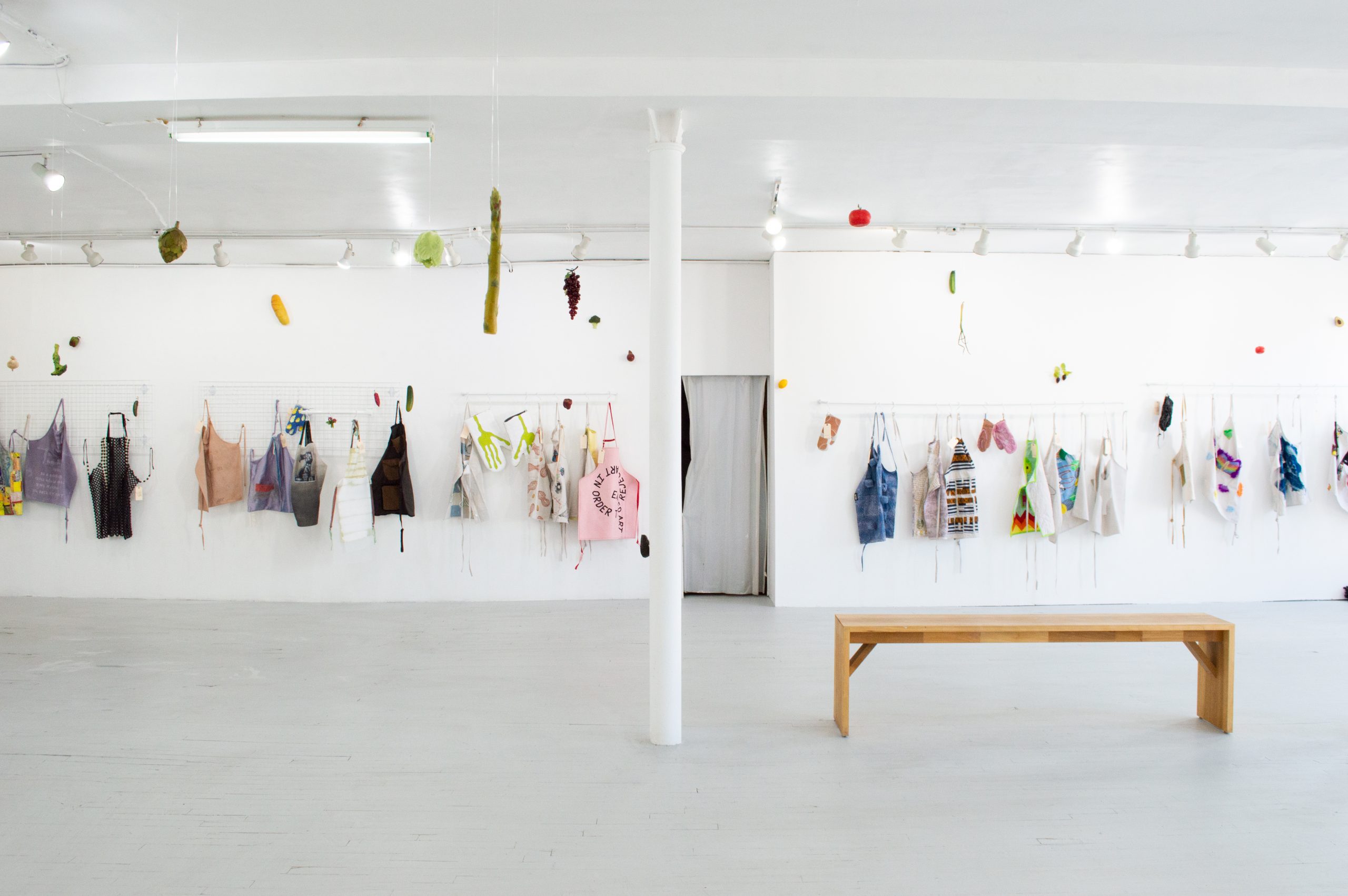 Featured image: Overview of LtdWear5 installed at the bright LVL3 gallery. The image is a landscape shot of about thirty colorful aprons and oven mitts displayed on racks on the far white wall of LVL3 gallery. In the midground of the shot is a long wooden bench and a white pole that cuts the gallery in half. Hanging from the ceiling are large plastic fruits and vegetables, including asparagus, grapes, and tomatoes. On the far wall, a large plastic baguette is hung on the wall as well offering a cheery, kitschy feeling to the gallery. Photo courtesy of LVL3.