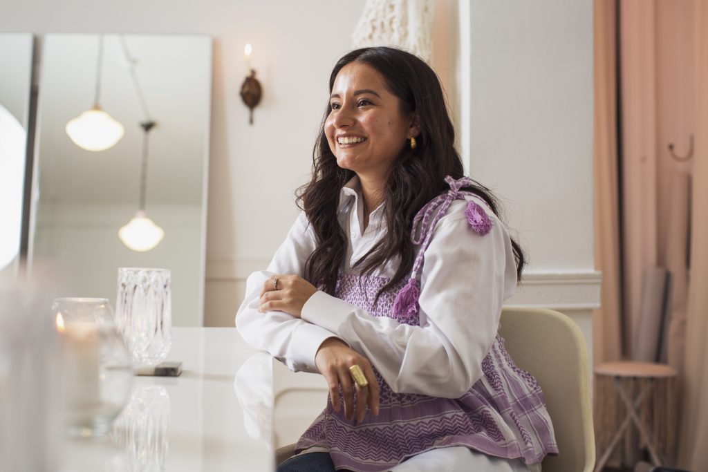 Image: Nelissa Carrillo is seated and smiling, facing away from the camera with her arms crossed and lightly resting on a reflective white table. The light casts a warm blush onto her face. The right side of the photo features peach lighting onto the dressing room of the showroom, and the left side of the photo is filled with a crystal glass, a candle, and the reflection of a pendant light in the large floor mirror. Lavender pompoms fall off of Carrillo’s shoulders onto her vintage white button down, and a gold statement ring on her right hand catches light from the pendant light. Photo by Kristie Kahns. 