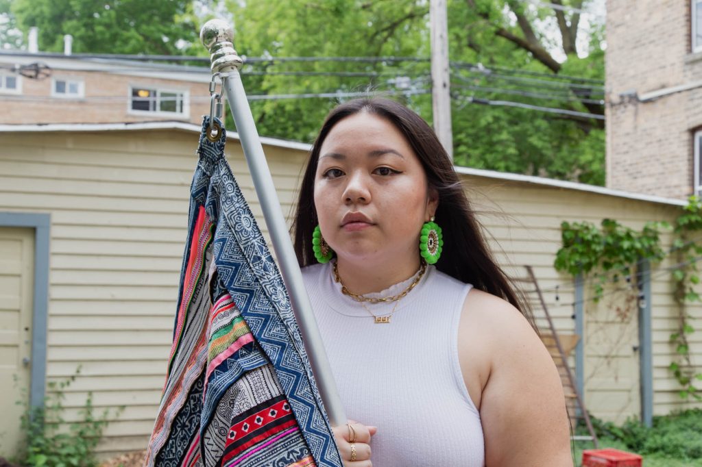 Image: Tshab Her holds her flag piece that was part of her performance We Are: Chang, Cheng, Chue, Fang, Hang, Her, Khang, Kong/Soung, Kue, Lee, Lor, Moua, Pha, Thao, Vang, Vue, Xiong, Yang while looking directly at the camera. She wears a white shirt, gold necklaces, and hand-made green earrings. Photo by Josh Johnson.