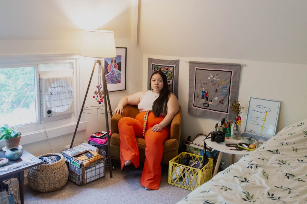 Image: A view of Tshab Her sitting in her room on an orange chair. She wears a white shirt and orange pants while looking directly at the camera. She sits amongst various containers of items. Two of her embroidery cloth pieces hang behind her. A lamp and a window are on the lefthand side of the frame. Photo by Josh Johnson.