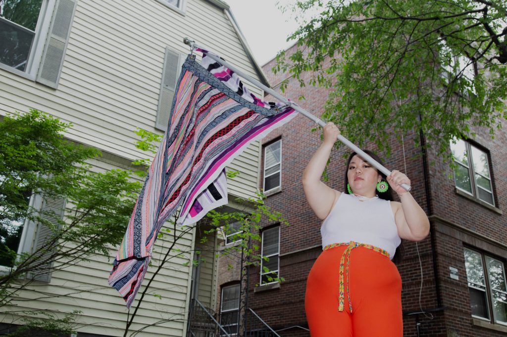 Image: Tshab Her waves her flag piece that was part of her performance We Are: Chang, Cheng, Chue, Fang, Hang, Her, Khang, Kong/Soung, Kue, Lee, Lor, Moua, Pha, Thao, Vang, Vue, Xiong, Yang while looking directly at the camera. She wears a white shirt, gold necklaces, orange pants, and hand-made green earrings. Photo by Josh Johnson.