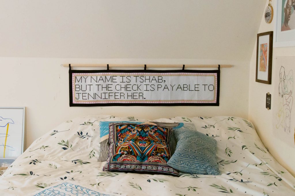 Image: A view of Tshab Her's bedroom, showing part of her bed and pillows. On the wall to the right, various artworks hang. Above the bed hangs an embroidery piece by Tshab Her, which reads: "MY NAME IS TSHAB, BUT THE CHECK IS PAYABLE TO JENNIFER HER." Photo by Josh Johnson.