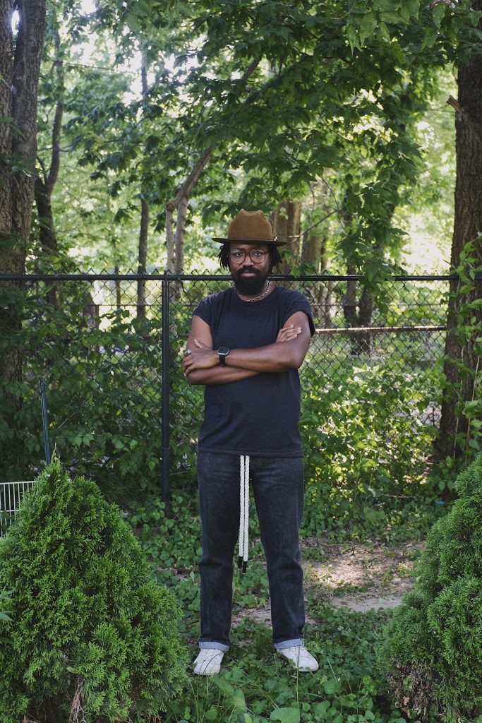 Image: A portrait of  Erick Minnis dressed in a dark top, jeans, a brown hat, and glasses, standing in the middle of greenery with arms folded. Photo by Erick Minnis.
