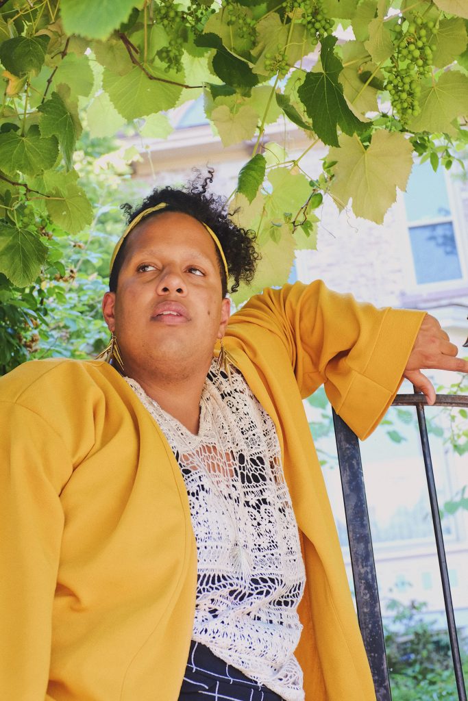 Image: A portrait of Alexander Martin dressed in a yellow jacket, white top, and dark striped shorts, arm resting on the top of a metal fence and standing under a grape vine. Photo by Erick Minnis.