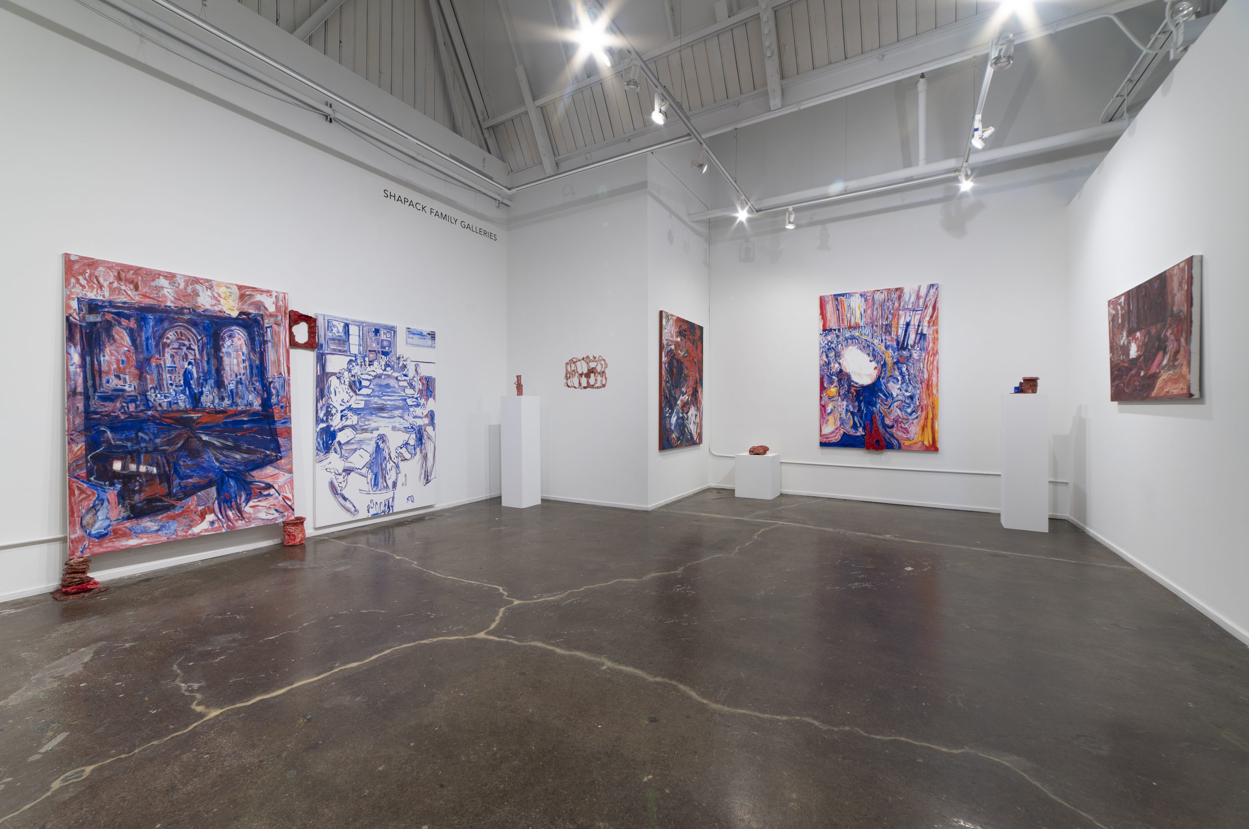Featured image: An installation view of Longing Compass at Chicago Artists Coalition, featuring the work of Karen Dana Cohen. The view of the gallery shows five paintings against white walls and four sculptural pieces with various additional three-dimensional pieces accompanying some of the canvases. The paintings all portray groups of people with large, gestural strokes of blue and red paint. Image courtesy of the artist.