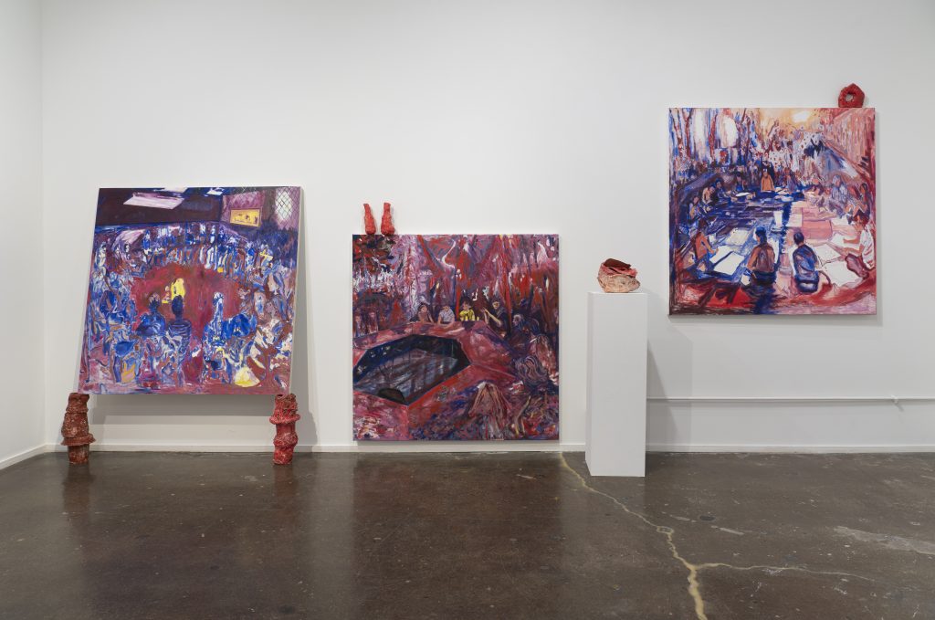 Image: An installation view of three paintings against a white wall with a three-dimensional piece sitting on a white pedestal to the right of the middle painting. The painting to the left uses large, gestural strokes in dark blues and reds to show a crowd of people standing in a circle. The painting in the middle shows a group of people sitting at a table with dark blues and maroon hues. The painting on the right is hung higher than the other two and shows a crowd of people in a room in reds and blues. Each painting has sculptures either on top or below the canvases. All work is by Karen Dana Cohen. Image courtesy of the artist.