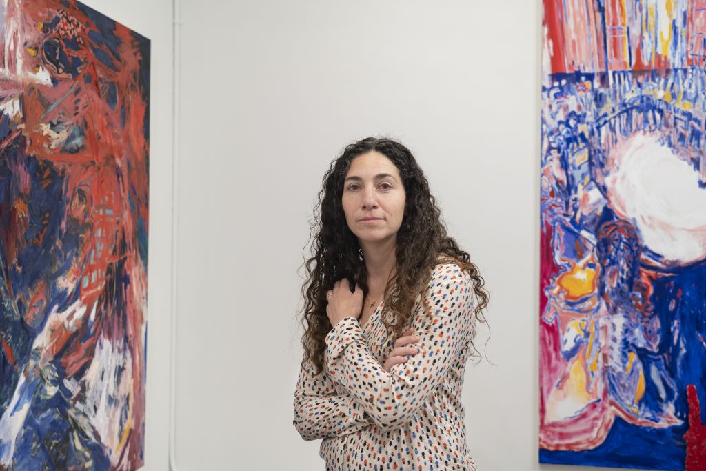 Image: The artist Karen Dana Cohen stands in front of and between two of her paintings looking directly at the viewer. Her arms are crossed and she has long brown hair. Image courtesy of the artist.