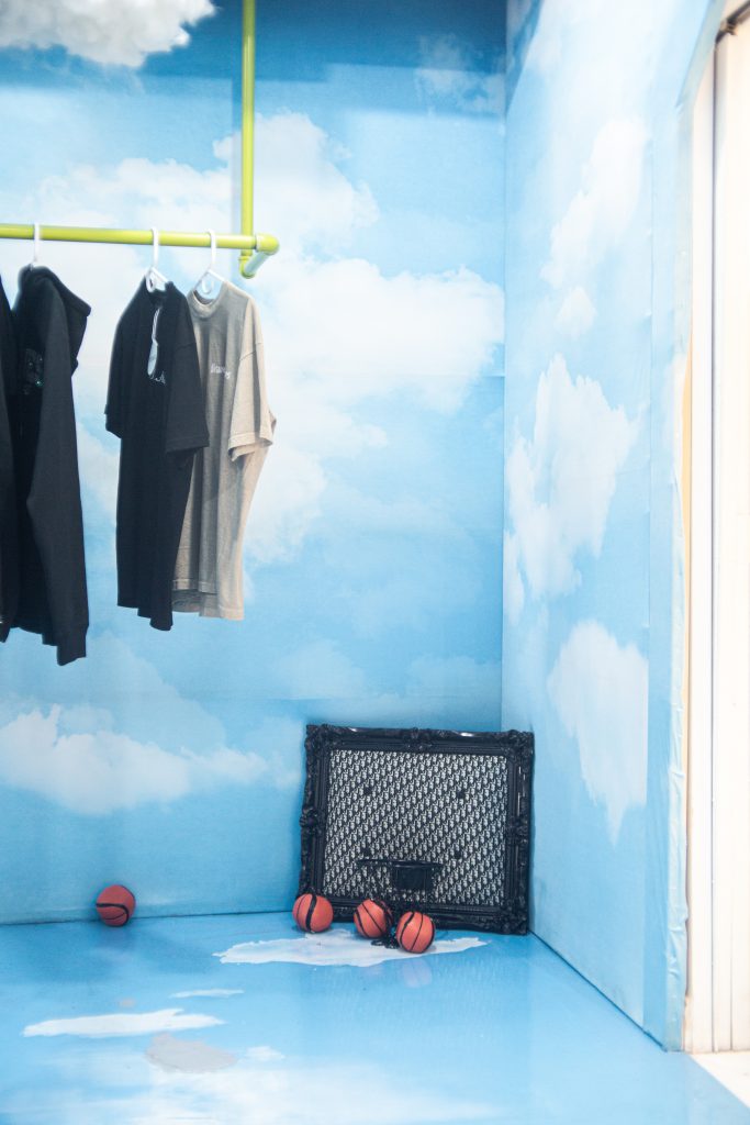 Image: A pop-up at Congruent Space featuring Hoop Dreams Studio. The walls and floor are covering by a blue sky and clouds. Basketballs and a basket and backboard sit on the ground. Clothes hang on a rack to the left. Photo by Dally Dew Drop.