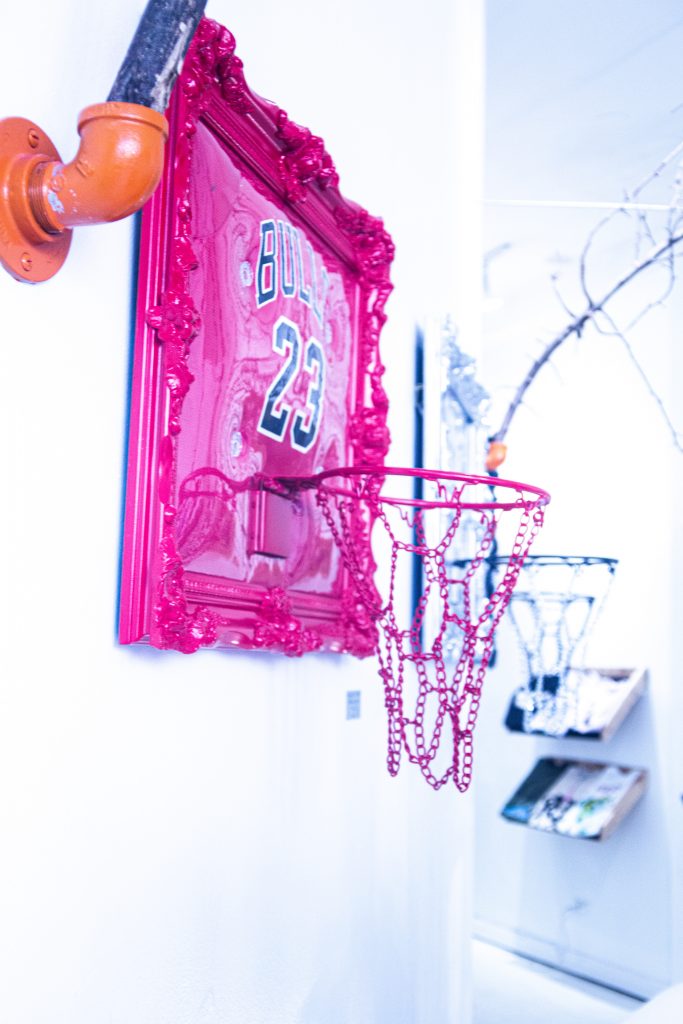 Image: A pop-up at Congruent Space featuring Hoop Dreams Studio. A magenta basketball hoop and headboard hangs on the white wall, with additional hoops in the background. Photo by Dally Dew Drop.