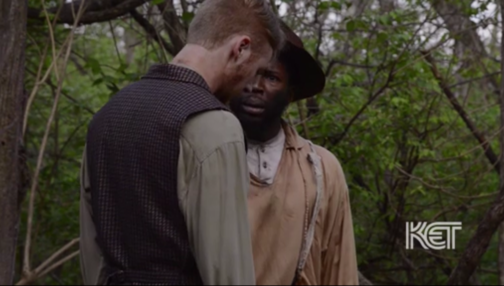 Image: A film still from The Categories Black and White. Actor Caleb Farley plays Archy, and actor Landon Horton plays Thomas. A man who appears Black talks to a man who appears white in a wooded area. Image courtesy of Mourning the Creation of Racial Categories Project.