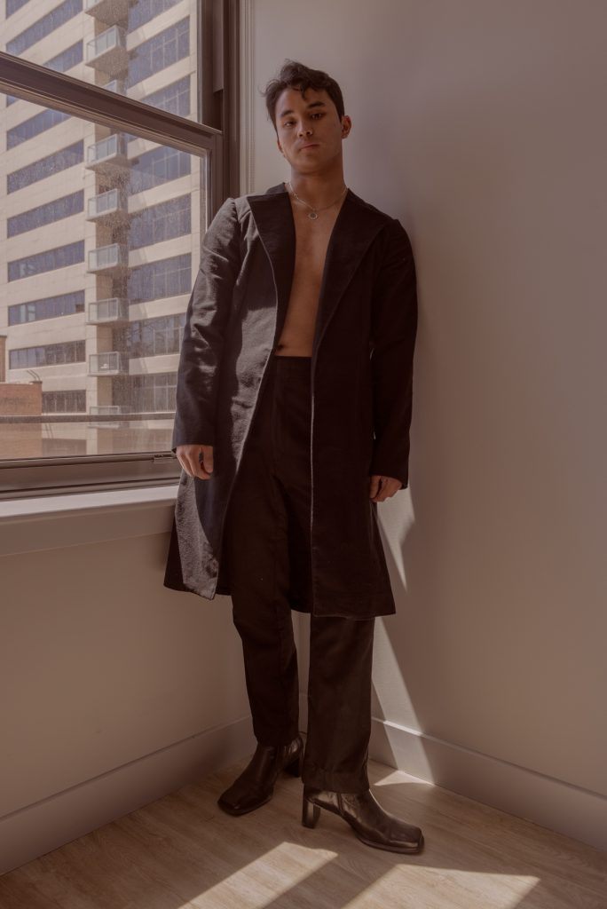 Image: Sam Fissell stands in front of a window while wearing a dark-colored, collared jacket. The jacket is open, revealing a silver neckless. The trousers are also dark and he is wearing black, healed shoes. Photo by Ryan Edmund Thiel.