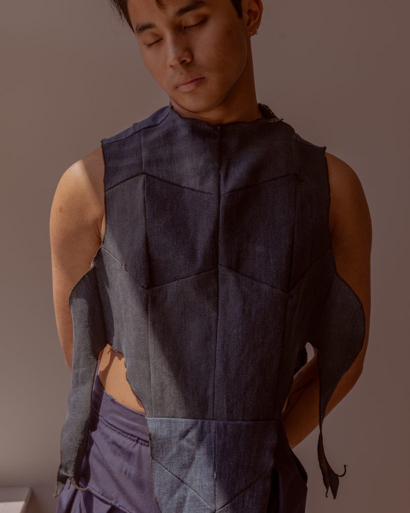 Image: Sam Fissell has one arm behind his back while looking down. He is wearing a dark-colored garment with ripped detailing and a geometric-like pattern. Photo by Ryan Edmund Thiel.