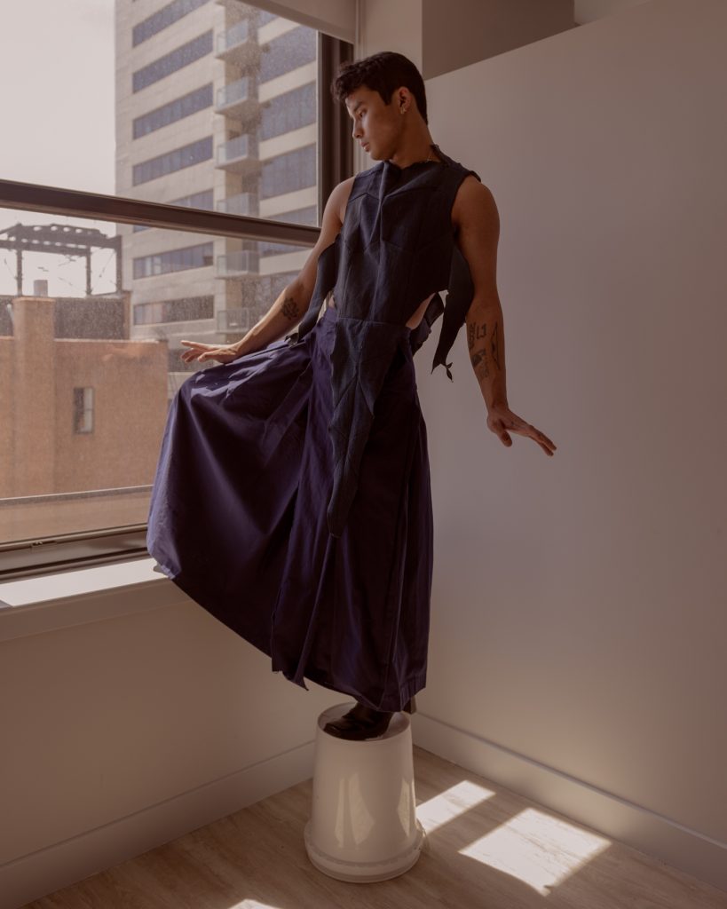 Image: Sam Fissell stands in front of a window and a white wall while posing with his arms slightly out and with one leg bent upwards while standing on a white bucket. He is wearing a dark, violet/blue garment that has ripped details towards the top and spreads out into a skirt shape towards the bottom. Photo by Ryan Edmund Thiel.