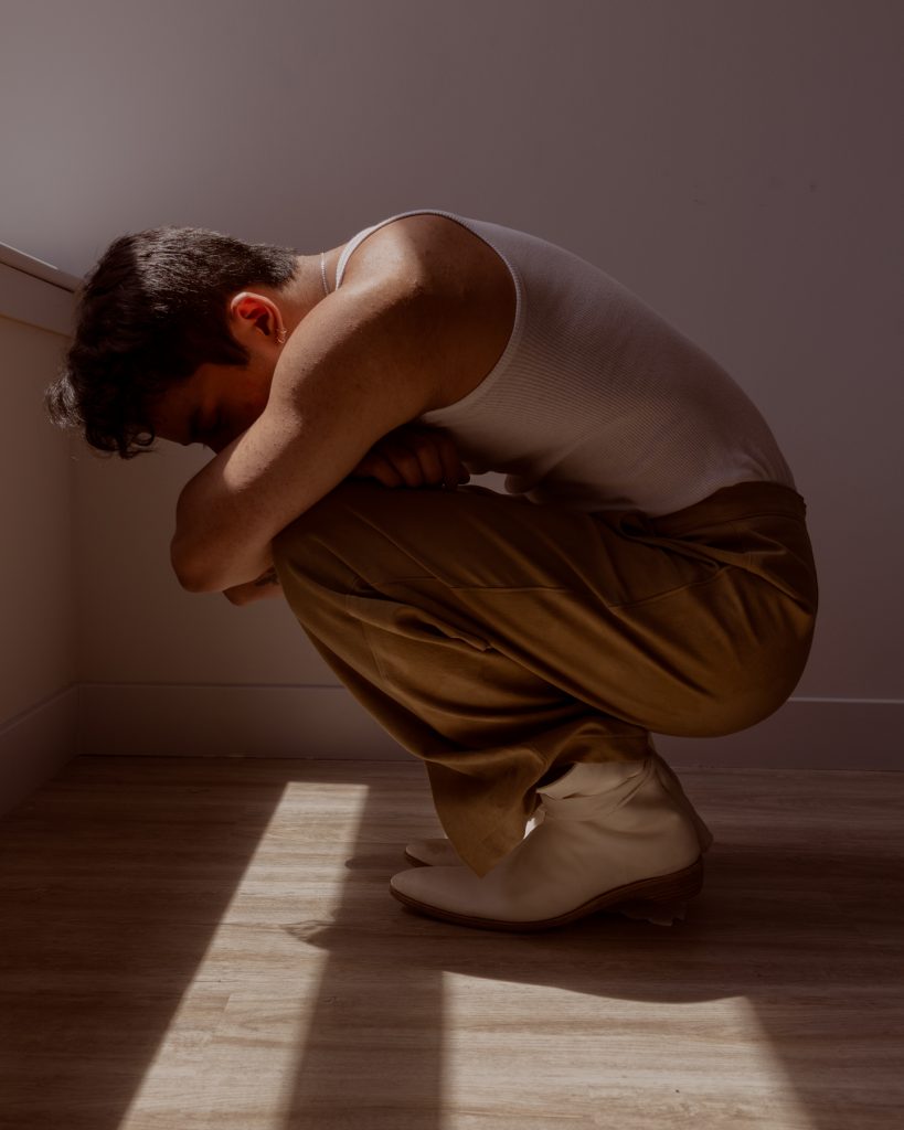 Image: Sam Fissell crouches while wearing tan trousers, a white tank top, a silver necklace, and white shoes with a white cloth tied around the ankle. Photo by Ryan Edmund Thiel.