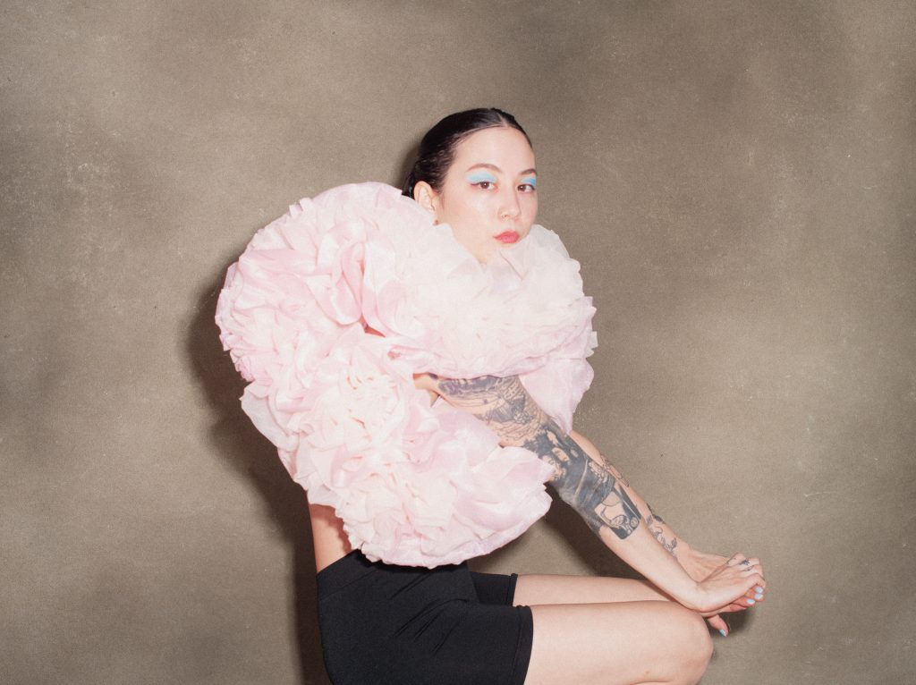 Image: A portrait of Michelle Zauner by Tonje Thilesen. Zauner sits while looking at the camera. She is wearing blue eye shadow, black shorts, and a pink, puffy top.