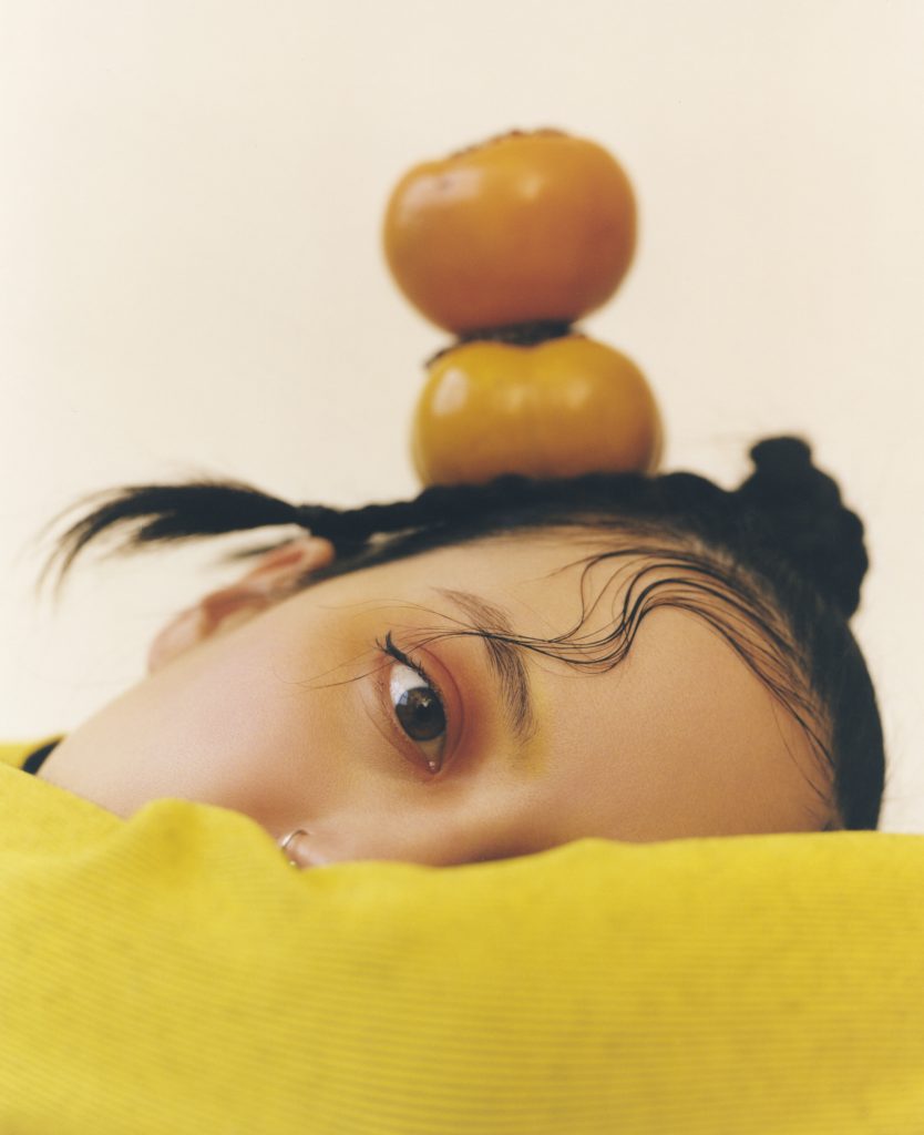 Image: A portrait of Michelle Zauner by Peter Ash Lee. Zauner faces the camera with her head on her arm. She wears yellow while wearing yellow and orange make up. An orange vegetable sits on her head.