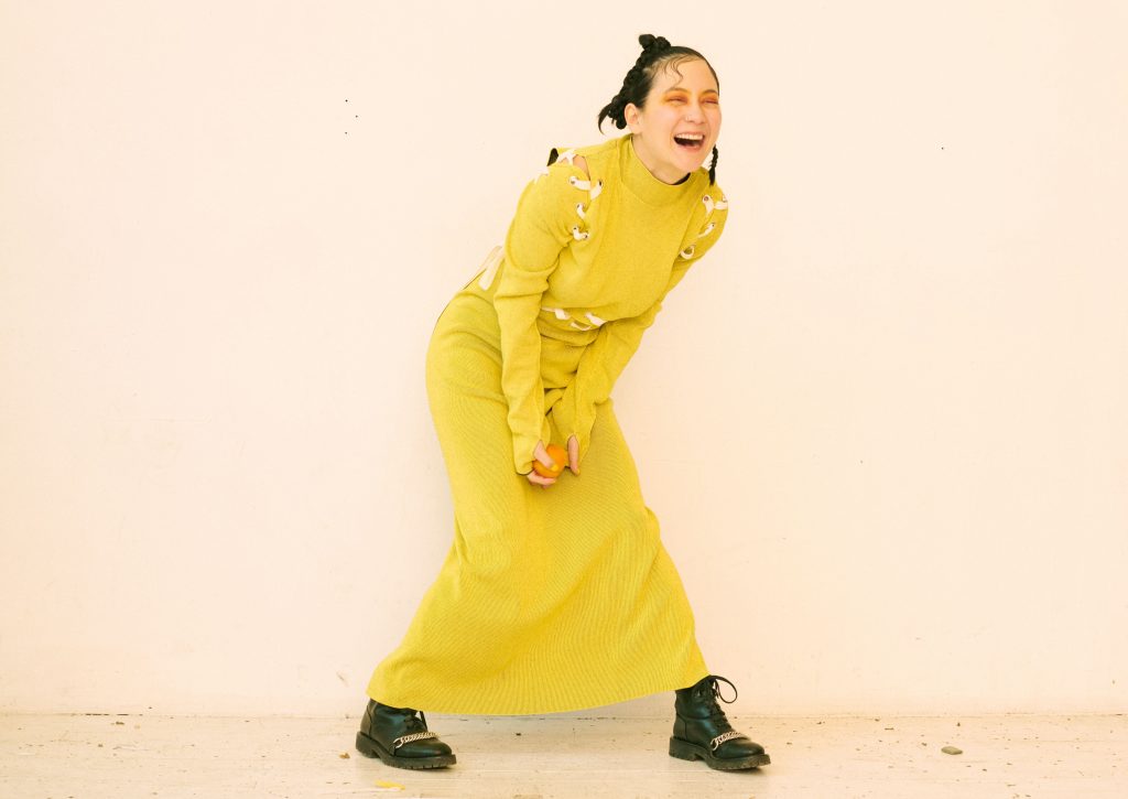 Image: A portrait of Michelle Zauner by Peter Ash Lee. Zander stands in front of a white wall while wearing a bright yellow dress and black boots. Her hair is in braids and she wears orange eye shadow while laughing.