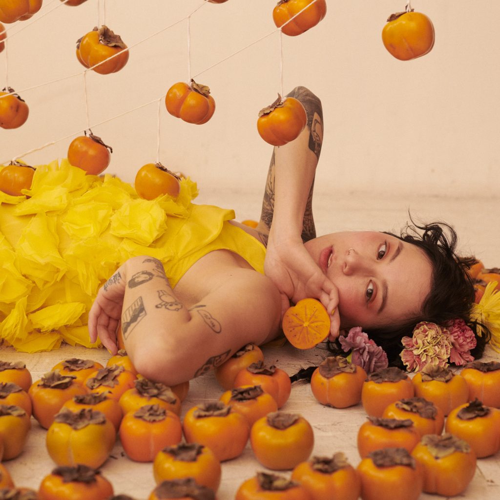 Image: A portrait of Michelle Zauner by Peter Ash Lee. Zander lays on the floor surrounded by orange vegetables suspended above her and laying around her. She holds one in her hand with flowers around her head.