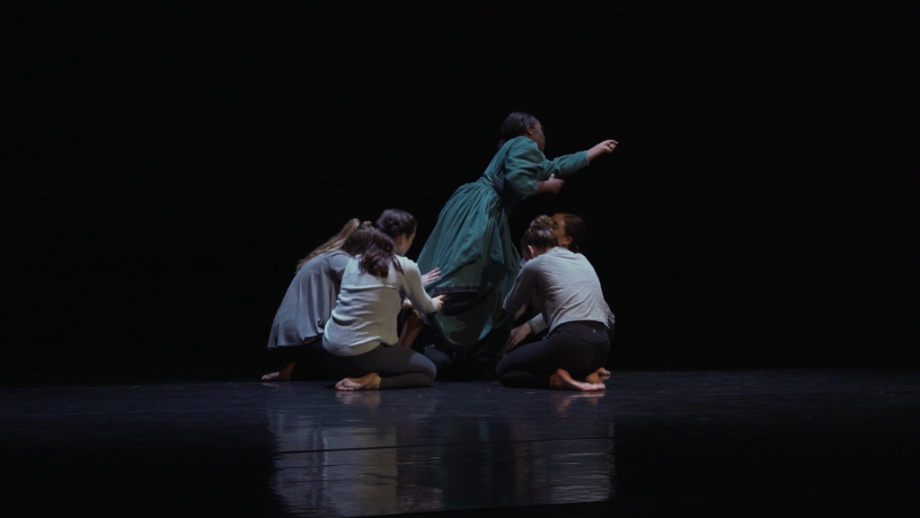 Image: A film still from Let Our Loss Be Heard. Actress Lavette Patterson as Margaret Garner is surrounded by ensemble dancers on stage while reaching out to our right. The background is completely dark. Courtesy of Mourning the Creation of Racial Categories Project.