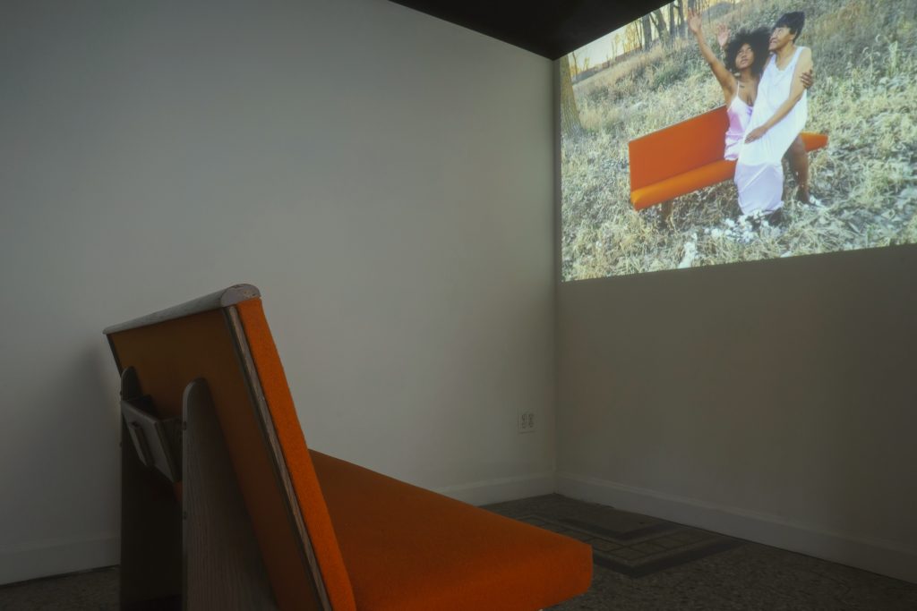 Image: A view of a video installation in the exhibition "Dreamscapes: Imaginings of a Black Pastoral" at Roots and Culture. The video shows two Black people sitting on an orange couch in in the grass. Photo by Colectivo Multipolar.