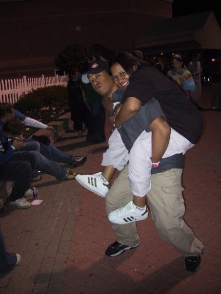 Image: Vertical outside photograph of Kay on the back of a POC male party go-er. Kay smiles at the camera, wearing eyeglasses, a black T-shirt, white cargo pants, white Adidas shell-toe sneakers with gold stripes. The man holding up Kay on his back is looking at the camera, wearing a grey T-shirt, khaki cargo pants, and black sneakers. They’re standing on beige and red brick pavement. There are other partygoers in the photo. To the left of them are three people sitting on an elevated portion of the pavement next to plants. Everyone else is behind Kay, unaware of the photo being taken. Photo courtesy of Kay.