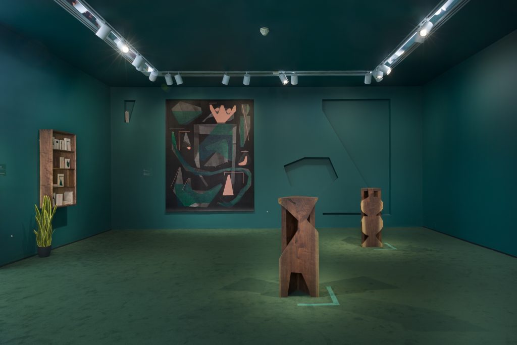 Image: An installation view of Victoria/Veronica: Making Room. The back of the room displays an abstract painting by Caroline Kent with an organic shape cut out of the wall to its right. The middle of the room contains two wooden sculptures. A shelf and a plant are displayed on the lefthand side. The entire room is deep green. Image courtesy of the artist and PATRON Gallery, Chicago. Photography by Evan Jenkins.