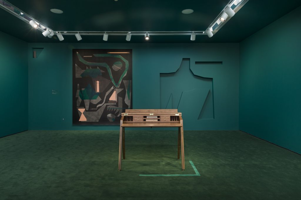 Image: An installation view of Victoria/Veronica: Making Room. The back of the room displays an abstract painting by Caroline Kent with an organic shape cut out of the wall to its right. The middle of the room contains a desk. The entire room is green. Image courtesy of the artist and PATRON Gallery, Chicago. Photography by Evan Jenkins.